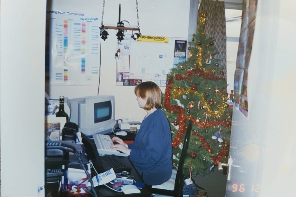 A young women sat at an old computer, on a messy desk. There is a Christmas tree behind her.